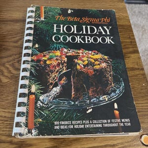 May include: A vintage spiral-bound cookbook titled "Holiday Cookbook" by The Beta Sigma Phi International. The cover features a festive cake on a glass plate, surrounded by candles. The book promises 900 recipes and ideas for holiday entertaining.