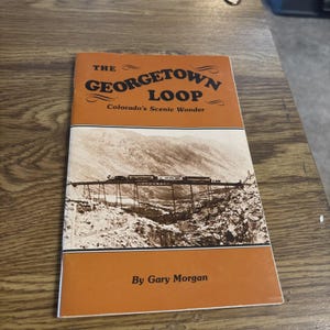May include: A vintage book titled "The Georgetown Loop" with a sepia-toned image of a train on a bridge in the mountains. The cover is orange and white with text that reads "Colorado's Scenic Wonder" and "By Gary Morgan."