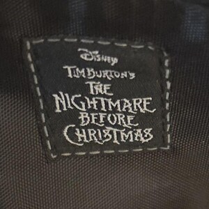 Puede incluir: Una etiqueta rectangular negra con texto blanco y un borde cosido. La etiqueta dice "Disney Tim Burton's The Nightmare Before Christmas". La etiqueta es probablemente de un producto relacionado con la pel&iacute;cula.