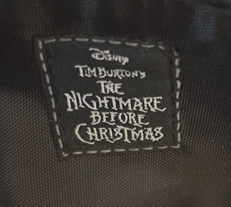 Puede incluir: Una etiqueta rectangular negra con texto blanco y un borde cosido. La etiqueta dice "Disney Tim Burton's The Nightmare Before Christmas". La etiqueta es probablemente de un producto relacionado con la pel&iacute;cula.