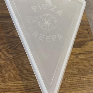 Op de afbeelding: Een doorschijnend, driehoekig plastic deksel voor een opbergbak met een opstaande rand. Het midden bevat een cirkelvormige afbeelding van een gesneden pizza met de tekst PIZZA KEEPA in reliëf in een halve cirkel boven en onder de afbeelding.