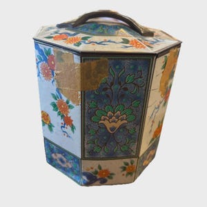 Könnte beinhalten: Eine antike, sechseckige Blechdose mit Griff. Die Dose ist mit Blumenmustern in Orange, Blau und Grün auf weißem Grund verziert. Deckel und Seiten weisen aufwendige Designs auf, wobei einige Bereiche Abnutzungserscheinungen aufweisen.