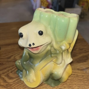 May include: A ceramic frog-shaped planter with a light green and yellow color scheme. The frog has large black eyes, a smiling mouth, and is holding a yellow coin. The planter is designed to resemble a tree stump.