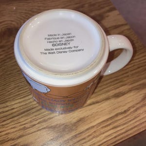 May include: A white ceramic mug with a handle, viewed from the bottom. The base features printed text: Made in Japan, Fabrique en Japon, Hecho en Japon, &copy;DISNEY, Made exclusively for The Walt Disney Company. The side shows partial colourful graphics.