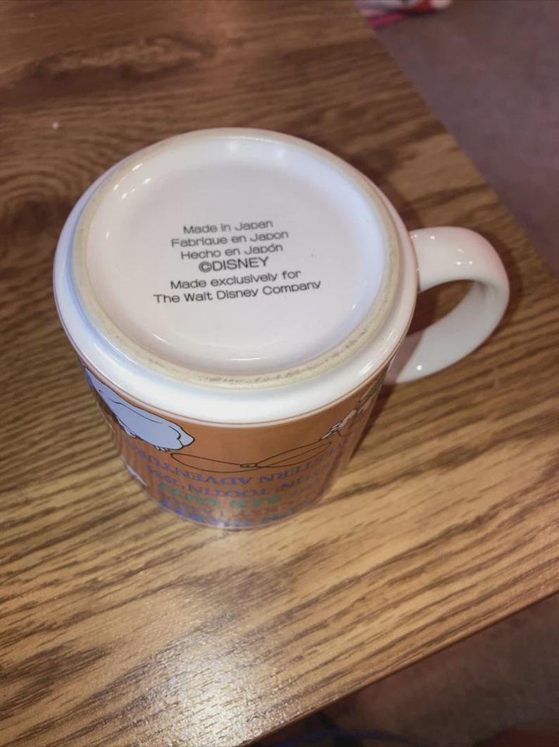 May include: A white ceramic mug with a handle, viewed from the bottom. The base features printed text: Made in Japan, Fabrique en Japon, Hecho en Japon, &copy;DISNEY, Made exclusively for The Walt Disney Company. The side shows partial colourful graphics.