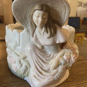 May include: A ceramic planter featuring a seated woman in a wide-brimmed hat, holding a bouquet of flowers. The planter is off-white with pink and green floral accents. The woman's dress is a soft pink.