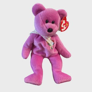 May include: A pink teddy bear with a black nose and eyes. It has a white ribbon around its neck with the text "2004" and a red heart-shaped tag attached to its ear. The bear is a classic soft toy.