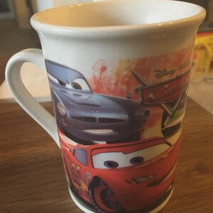 May include: White ceramic mug featuring characters from the Disney Pixar Cars film. The mug has a handle and displays images of Lightning McQueen and other race cars. The mug is cylindrical and suitable for hot or cold drinks.