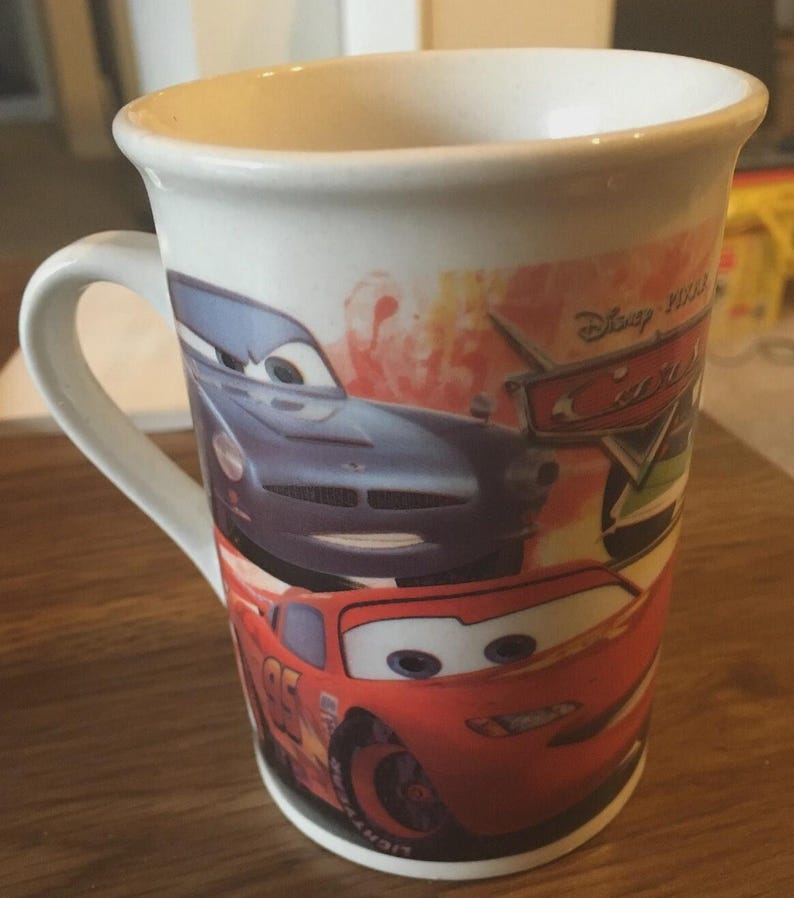 May include: White ceramic mug featuring characters from the Disney Pixar Cars film. The mug has a handle and displays images of Lightning McQueen and other race cars. The mug is cylindrical and suitable for hot or cold drinks.