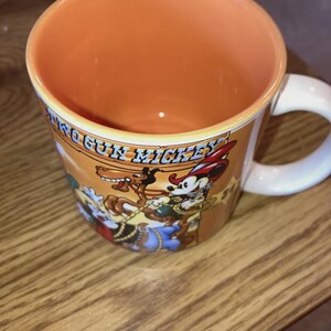 May include: A ceramic mug with a light orange interior and a white handle. The exterior features a graphic of Two Gun Mickey with a horse and the text Two Gun Mickey printed in blue. The mug has a warm orange background and a white handle.