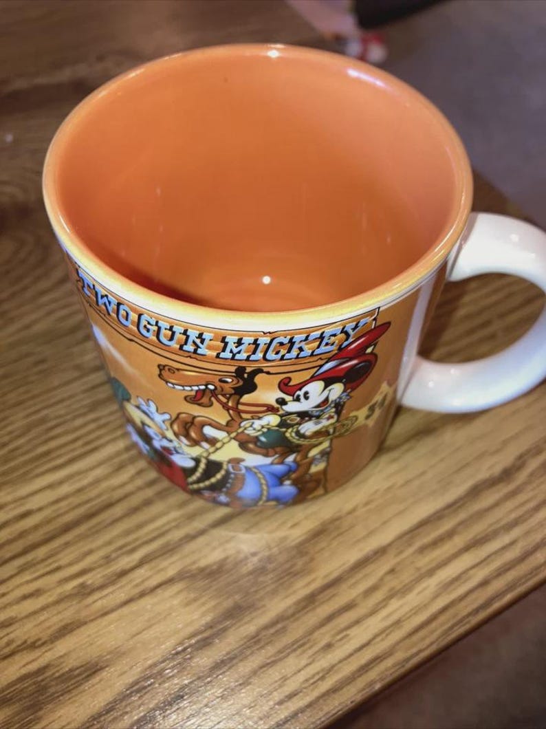 May include: A ceramic mug with a light orange interior and a white handle. The exterior features a graphic of Two Gun Mickey with a horse and the text Two Gun Mickey printed in blue. The mug has a warm orange background and a white handle.