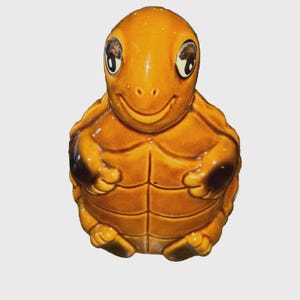 Puede incluir: Figura de tortuga de cerámica con acabado marrón anaranjado brillante. La tortuga tiene ojos grandes y expresivos, boca sonriente y caparazón detallado. Artículo decorativo.