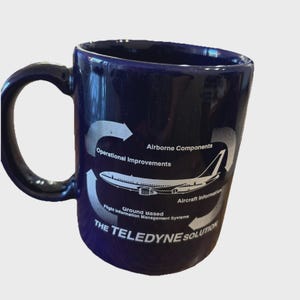 May include: A dark blue ceramic mug featuring a white graphic of an airplane surrounded by arrows and text including Airborne Components, Operational Improvements, Aircraft Information, Ground Based Flight Information Management Systems, and THE TELEDYNE SOLUTION.