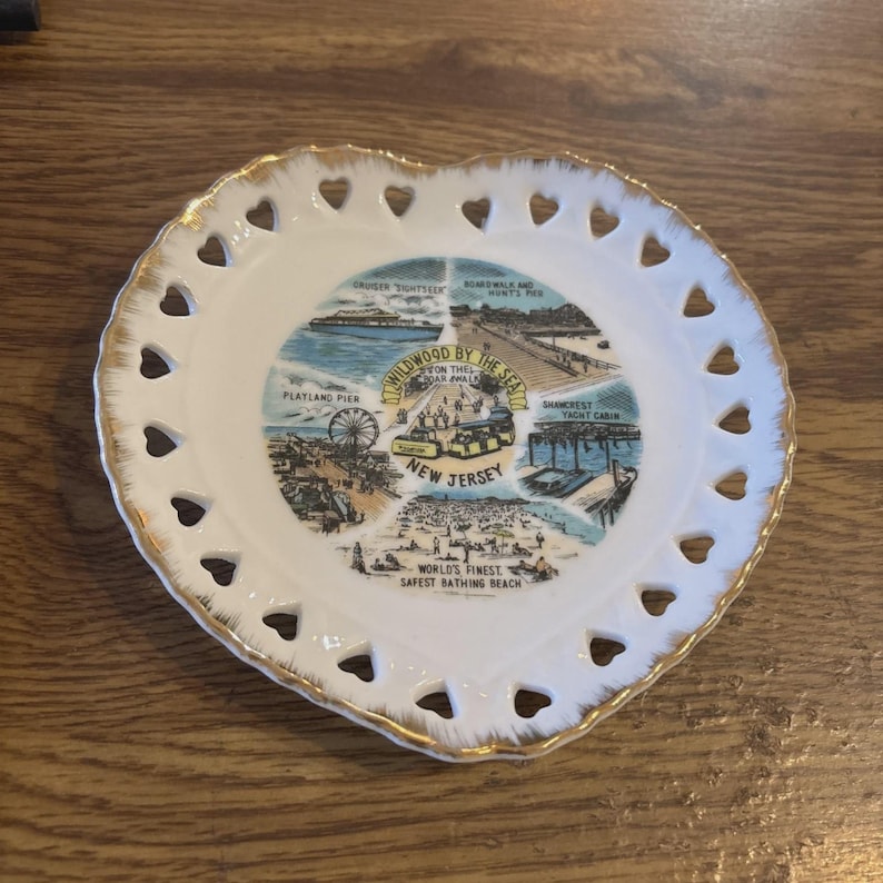 May include: Heart-shaped decorative plate with a gold rim and heart-shaped cutouts. The center features a colorful illustration of Wildwood, New Jersey, with text that reads "Wildwood by the Sea" and "New Jersey."