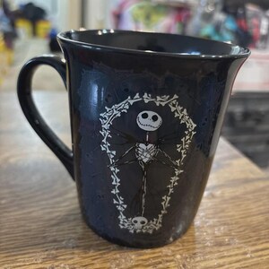 May include: Black ceramic mug featuring a white illustration of Jack Skellington from The Nightmare Before Christmas. The design includes a coffin-shaped frame and skull accents. The mug has a curved handle.