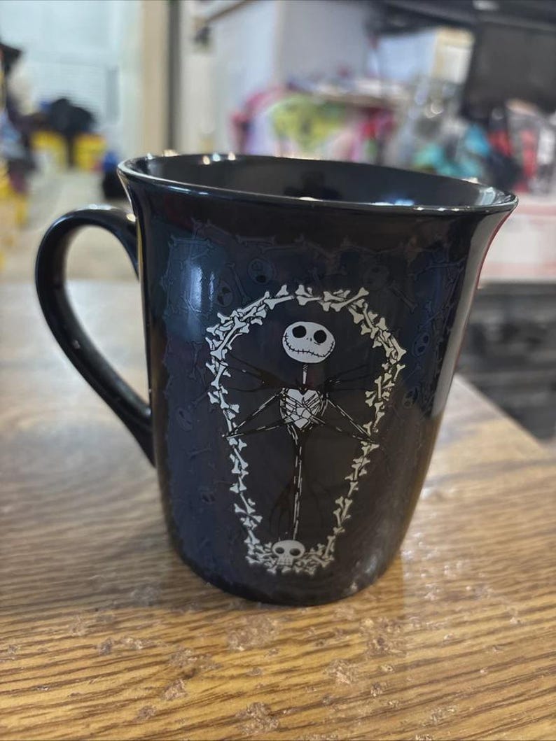 May include: Black ceramic mug featuring a white illustration of Jack Skellington from The Nightmare Before Christmas. The design includes a coffin-shaped frame and skull accents. The mug has a curved handle.