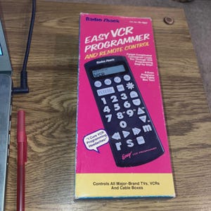 May include: A pink and black Radio Shack Easy VCR Programmer and Remote Control box. The box features the text "EASY VCR PROGRAMMER AND REMOTE CONTROL" and "Controls All Major-Brand TVs, VCRs And Cable Boxes."