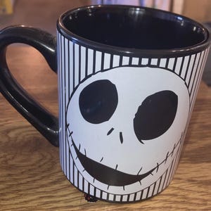May include: Black and white ceramic mug featuring a smiling face design. The mug has a black handle and interior, with vertical stripes and a character face on the exterior. A decorative item for fans of the character.