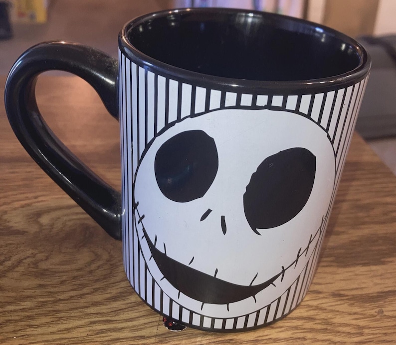 May include: Black and white ceramic mug featuring a smiling face design. The mug has a black handle and interior, with vertical stripes and a character face on the exterior. A decorative item for fans of the character.