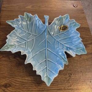 Könnte beinhalten: Eine keramikförmige Blattschale in Blau- und Grüntönen. Die Schale hat detaillierte Adern und eine glänzende Oberfläche. Ein goldener Aufkleber ist auf der Schale sichtbar. Die Schale befindet sich auf einer Holzoberfläche.