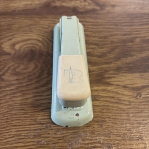 May include: Vintage mint green stapler with a cream-colored top. The stapler has a rectangular shape with rounded edges and a visible logo on the top. The stapler is placed on a wooden surface.
