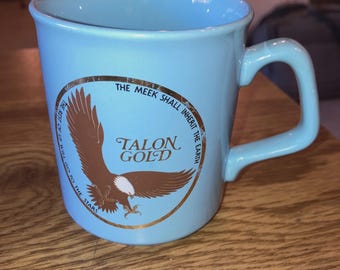 Może przedstawiać: Jasnoniebieski ceramiczny kubek z zakrzywionym uchwytem. Kubek posiada złoty emblemat z brązowym orłem i napisem "TALON GOLD". Na kubku wydrukowano również tekst "THE MEEK SHALL INHERIT THE EARTH" i "THE REST OF US WILL GO TO THE STARS".