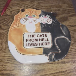 May include: A ceramic cat-shaped dish with a calico cat on the left and a black cat on the right. The dish has a white rectangular sign that reads "THE CATS FROM HELL LIVES HERE". The cats have green eyes and pink noses.