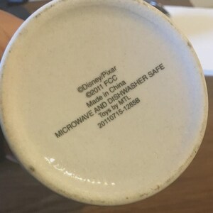 May include: The base of a white ceramic mug with text that reads "&copy;Disney/Pixar &copy;2011 FCC Made in China MICROWAVE AND DISHWASHER SAFE Toys by MTL 20110715-1285B". The mug is slightly off-white.