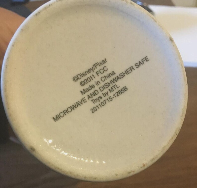 May include: The base of a white ceramic mug with text that reads "&copy;Disney/Pixar &copy;2011 FCC Made in China MICROWAVE AND DISHWASHER SAFE Toys by MTL 20110715-1285B". The mug is slightly off-white.