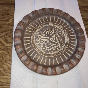 Peut inclure: Une assiette en céramique marron avec un bord festonné en relief. Le centre présente un motif détaillé de couleur crème représentant une figure stylisée dans un motif circulaire. L'assiette repose sur une serviette en papier blanche.