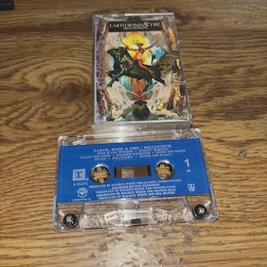 Peut inclure: Une cassette audio vintage de l'album "Millennium" d'Earth, Wind & Fire. Le boîtier de la cassette présente une illustration de l'album avec une personne chevauchant un taureau noir. La cassette bleue porte le titre des chansons en blanc.