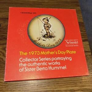 Può includere: Un piatto quadrato rosso con un'illustrazione circolare di un bambino che pesca, con la scritta "Mother's Day 1973". Il piatto fa parte della serie Collector che ritrae le opere di Suor Berta Hummel.