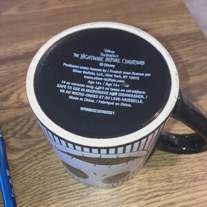 May include: A black and white ceramic mug featuring a design from Tim Burton's "The Nightmare Before Christmas." The mug has a black handle and a white skull pattern around the base. The bottom of the mug has text including the Disney logo and product information.