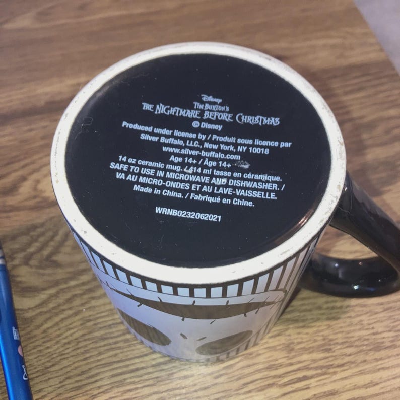 May include: A black and white ceramic mug featuring a design from Tim Burton's "The Nightmare Before Christmas." The mug has a black handle and a white skull pattern around the base. The bottom of the mug has text including the Disney logo and product information.