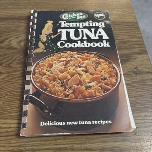 May include: A vintage cookbook titled "Tempting Tuna Cookbook" by Chicken of the Sea. The cover features a mermaid illustration and a photo of a tuna dish. The cookbook is spiral-bound and has the text "Delicious new tuna recipes".
