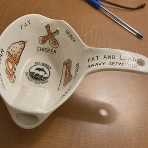 Puede incluir: Un separador de salsa de cerámica blanca con asa y boquilla. El cuenco está decorado con ilustraciones de jamón, pollo y bistec, con las palabras "FAT AND LEAN GRAVY SEPARATOR" impresas en el asa. También es visible el logotipo de Sea World.