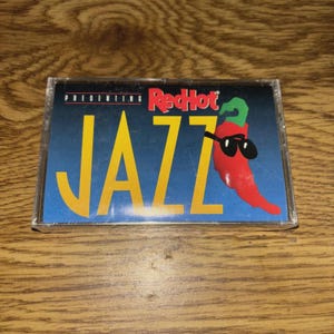 May include: A vintage cassette tape with a blue and yellow cover. The cover features the text "PRESENTING RedHot JAZZ" with a cartoon chili pepper wearing sunglasses. The cassette is in a clear plastic case.