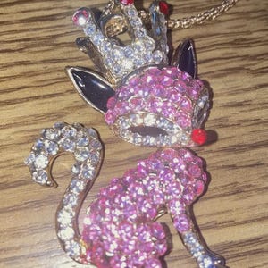 Può includere: Una collana color oro con un ciondolo a forma di volpe tempestata di gioielli con una corona. La volpe è ricoperta di strass rosa, con una coda di strass bianchi. La corona ha strass trasparenti e rossi. La collana è su una superficie di legno.