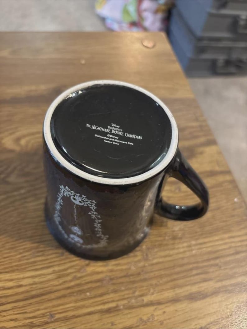 May include: Black ceramic mug featuring a design from "The Nightmare Before Christmas." The mug has a white rim and handle. The bottom of the mug has text that reads "The Nightmare Before Christmas" and "Dishwasher and Microwave Safe."