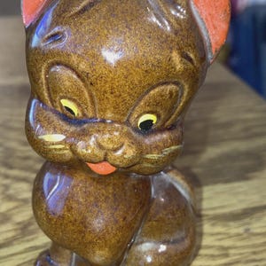 May include: A vintage ceramic cat figurine in shades of brown with orange accents on the ears and a red mouth. The cat has yellow eyes and is in a seated position. This collectible cat figurine is a decorative item.