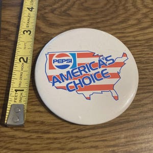 May include: A white, round Pepsi button with the text "AMERICA'S CHOICE" in red and blue, overlaid on a map of the United States. The button is approximately 2 inches in diameter.