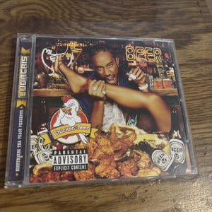 May include: A Ludacris Chicken-n-Beer music CD in a clear plastic jewel case. The cover art features a person holding a leg, a cartoon chicken logo, fried chicken, beer bottles, and the text Parental Advisory Explicit Content.
