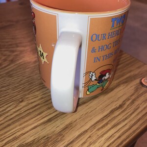 May include: A ceramic mug with an orange interior and exterior featuring a cartoon character wearing a red hat and green trousers. The text reads TWO OF OUR HEROES & HOG TIES IN THIS ROUNDUP. A white handle is attached to the side of the orange mug.