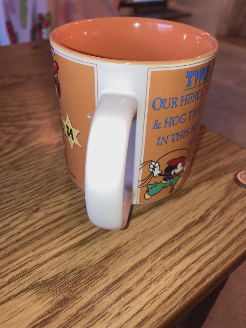 May include: A ceramic mug with an orange interior and exterior featuring a cartoon character wearing a red hat and green trousers. The text reads TWO OF OUR HEROES & HOG TIES IN THIS ROUNDUP. A white handle is attached to the side of the orange mug.