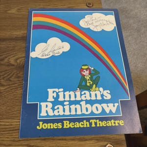 May include: A vintage Finian's Rainbow playbill with a blue background, a rainbow, and a cartoon leprechaun. The playbill has signatures and the text "Finian's Rainbow" and "Jones Beach Theatre".
