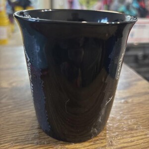 May include: A black ceramic cup with a flared rim. The cup has a glossy finish and appears to be empty. The cup is sitting on a wooden surface.