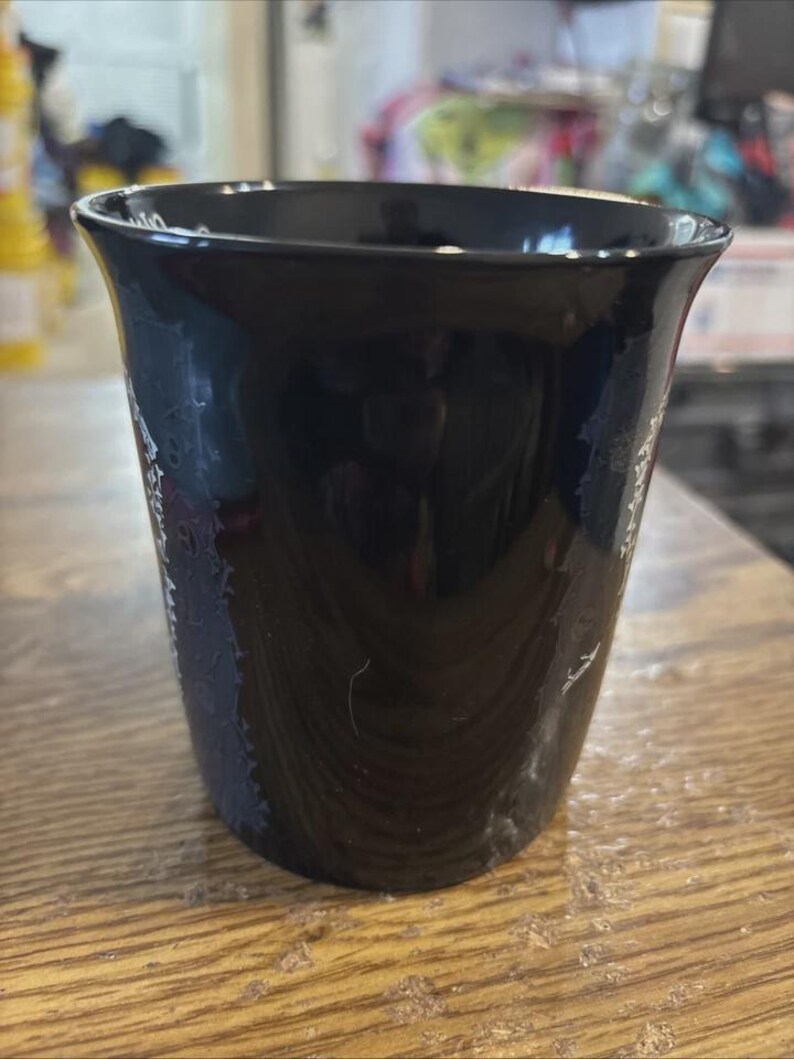 May include: A black ceramic cup with a flared rim. The cup has a glossy finish and appears to be empty. The cup is sitting on a wooden surface.