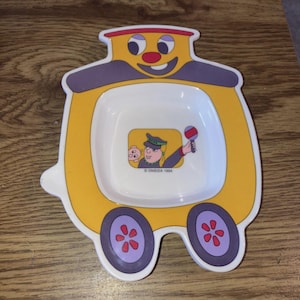 Puede incluir: Plato con forma de tren amarillo y morado, con una cara sonriente y ruedas. El interior blanco presenta una ilustración de dibujos animados de un conductor y un cerdo. El plato está marcado "© ONEIDA 1994".