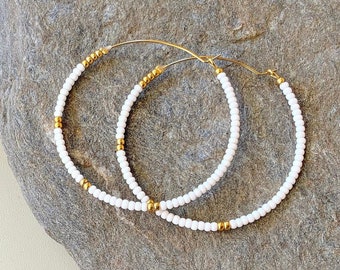 May include: A pair of gold hoop earrings with white beads. The earrings are on a gray rock.