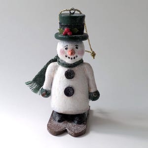 May include: A snowman ornament with a green top hat, scarf, and mittens. The snowman has a carrot nose, black button eyes, and a coal mouth. It stands on brown skis and has a gold string for hanging. A festive holiday decoration.